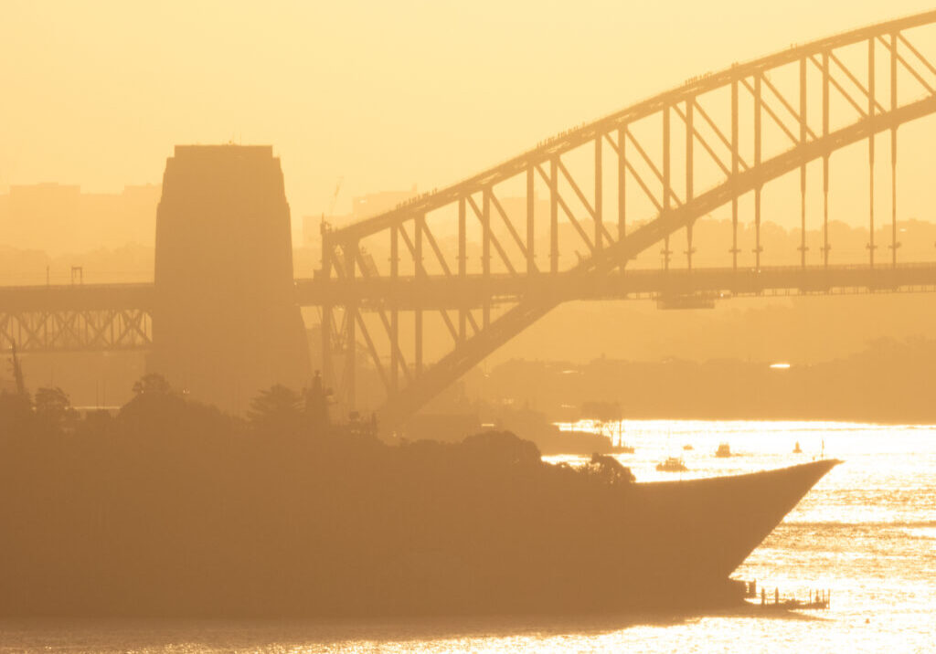 The sun is setting over Sydney Harbour. The curved flight deck bow of HMAS Adelaide and mast of HMAS Brisbane are visible, with the rest of the vessels hidden behind trees. docked at Garden Island naval base. Groups of people are visible climbing the Sydney Harbour Bridge. This image was taken from Dudley Page Reserve, Dover Heights in the eastern suburbs of Sydney on a hot and sunny afternoon shortly before sunset on 18 April 2025.