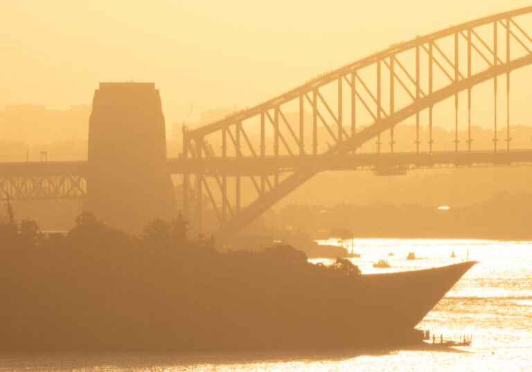 The sun is setting over Sydney Harbour. The curved flight deck bow of HMAS Adelaide and mast of HMAS Brisbane are visible, with the rest of the vessels hidden behind trees. docked at Garden Island naval base. Groups of people are visible climbing the Sydney Harbour Bridge. This image was taken from Dudley Page Reserve, Dover Heights in the eastern suburbs of Sydney on a hot and sunny afternoon shortly before sunset on 18 April 2025.